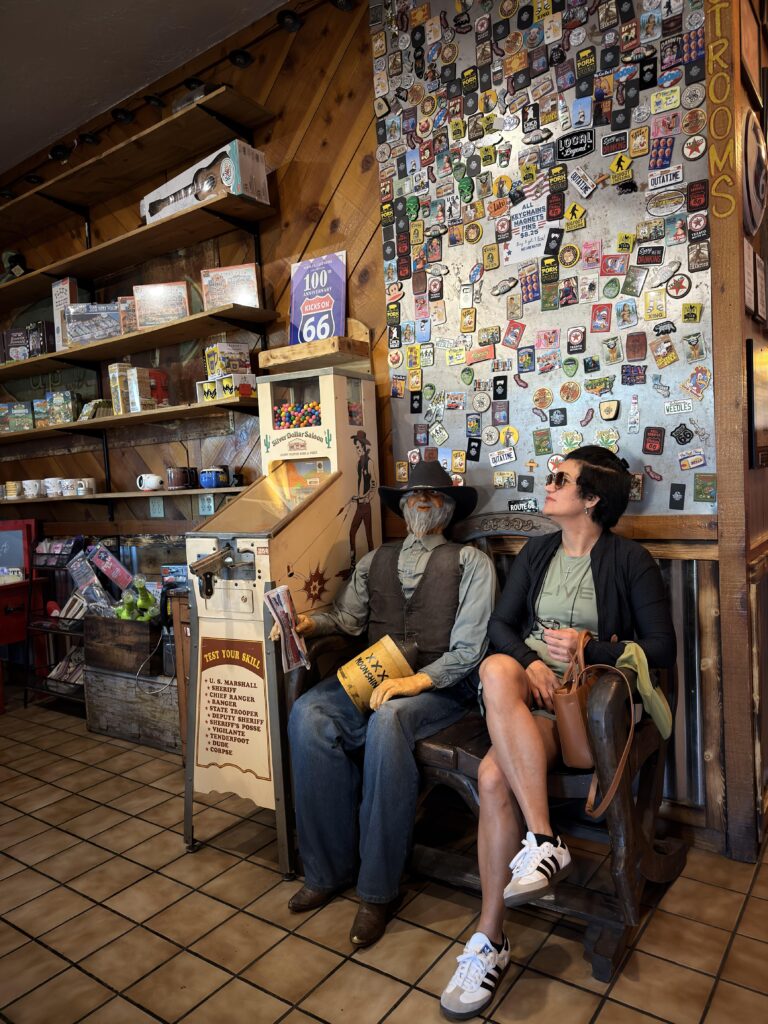 Frau sitzt neben lebensgroßer Cowboy-Figur in Route-66-Souvenirshop mit Magneten-Wand