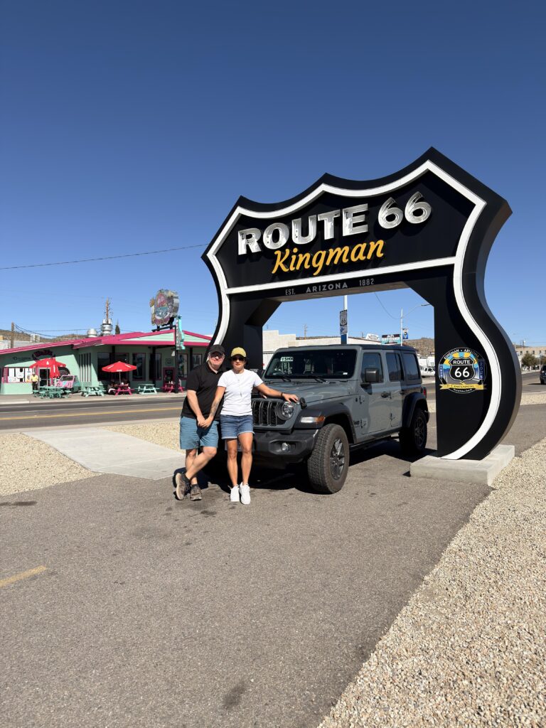 Paar vor dem großen Route 66 Kingman Arizona Schild mit grauem Jeep Wrangler