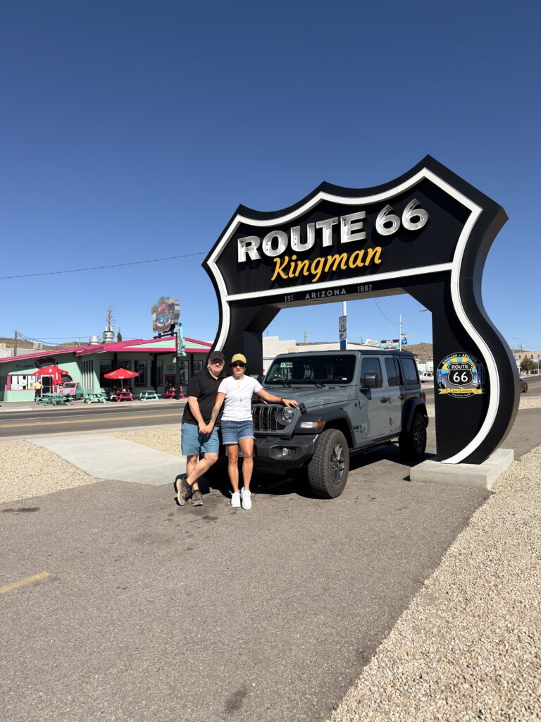 Mann und Frau stehen vor einem großen Route-66-Schild mit Aufschrift „Kingman, Arizona, Est. 1882", daneben ein grauer Jeep Wrangler, im Hintergrund ein buntes Diner