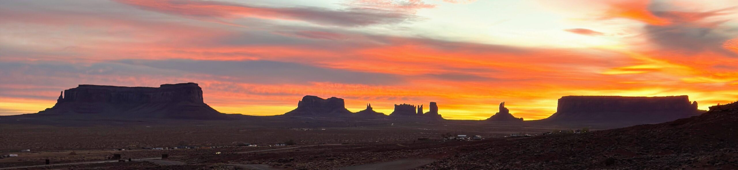 Breites Panoramafoto des Monument Valley bei Sonnenaufgang, alle Buttes als schwarze Silhouetten vor einem glutroten Himmel mit orangefarbenen Wolken.