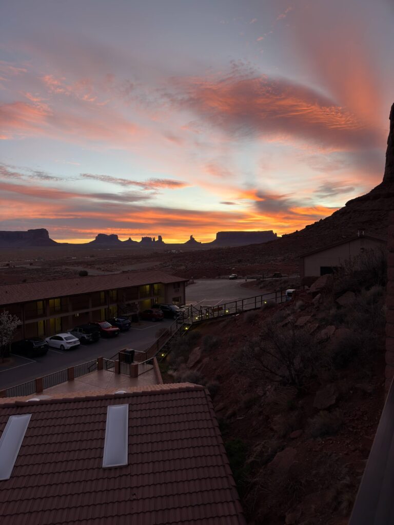 Orangeroter Sonnenaufgang über dem Monument Valley mit Buttes als Silhouetten, im Vordergrund Dach und Parkplatz der Goulding's Lodge.