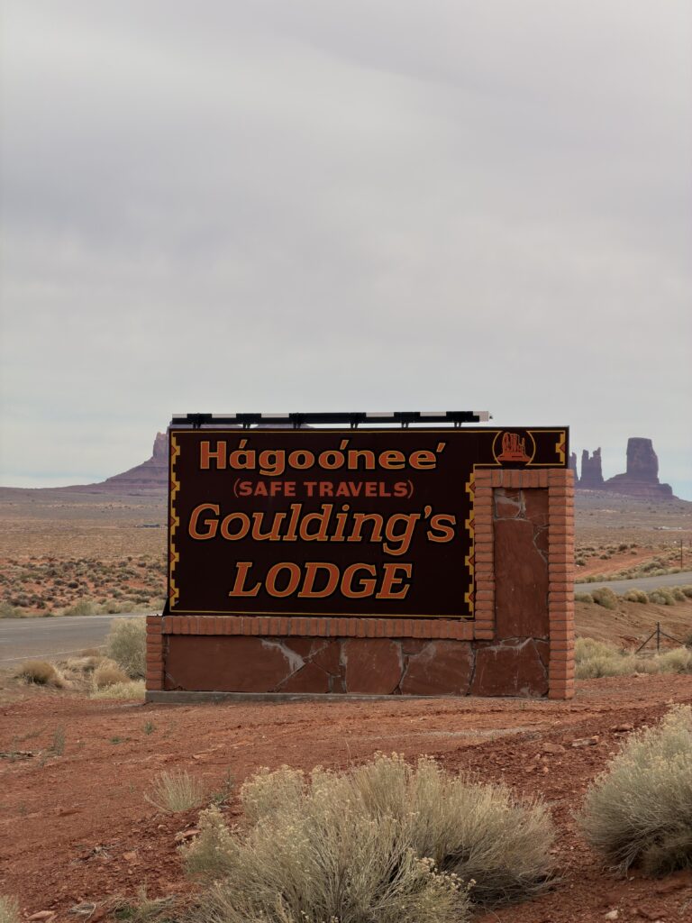 Eingangsschild der Goulding's Lodge mit dem Navajo-Wort „Hágoo'nee'" und der Übersetzung „Safe Travels", im Hintergrund Wüste und Buttes.
