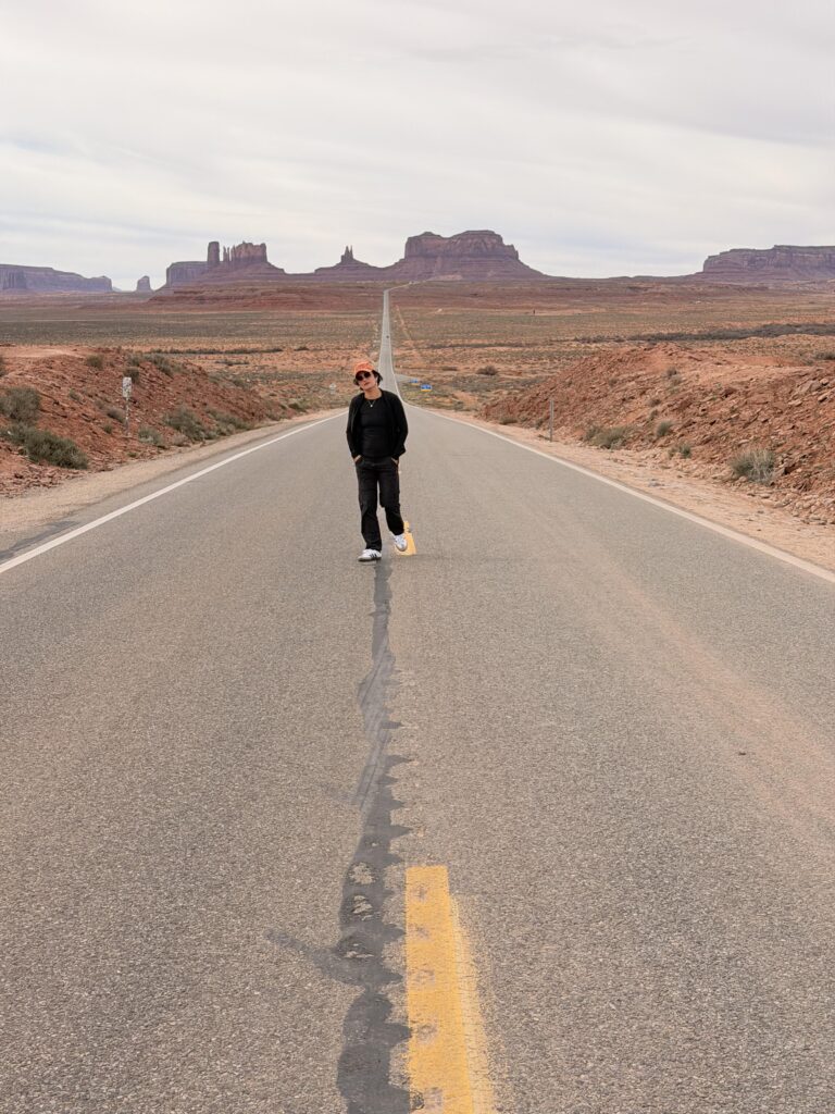 Frau mit orangem Cap steht frontal auf der Mittellinie des leeren Highway 163, hinter ihr die ikonischen Buttes des Monument Valley.