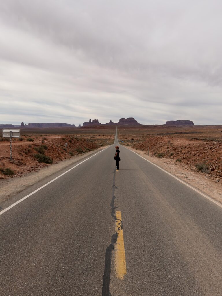 Frau mit orangem Cap steht seitlich auf der Mittellinie des leeren Highway 163, Buttes des Monument Valley als Silhouetten am Horizont.