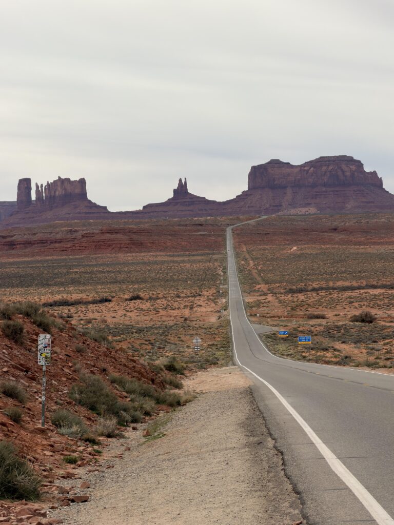 Leerer Highway 163 führt schnurgerade auf die Buttes des Monument Valley zu, links ein mit Stickern beklebter Pfosten, Wüstenlandschaft ringsum.