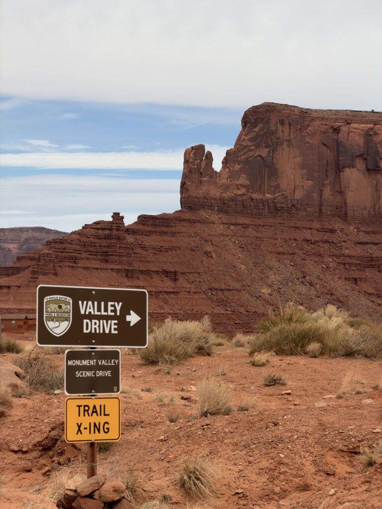 Braunes Schild „Valley Drive / Monument Valley Scenic Drive" mit gelbem „Trail X-ing" Schild davor, dahinter eine rote Sandsteinformation.