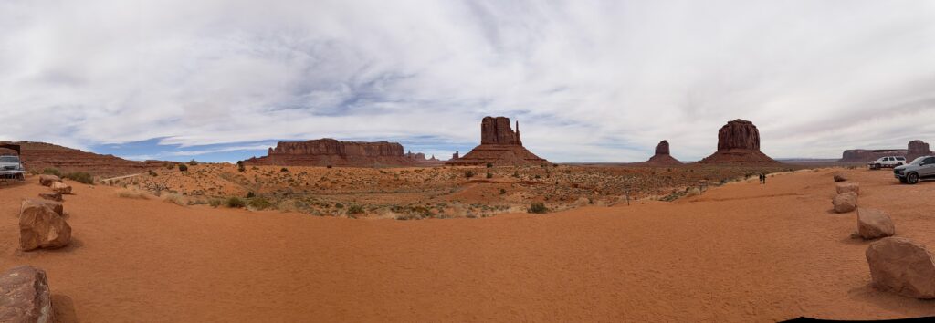 Breites Panoramafoto des Monument Valley mit West Mitten, East Mitten und Merrick Butte auf leuchtend rotem Sandboden unter bewölktem Himmel.
