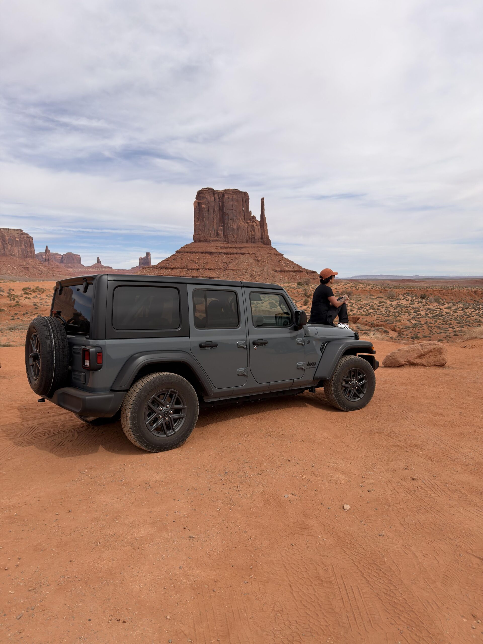 Grauer Jeep Wrangler steht auf rotem Sand im Monument Valley, daneben sitzt eine Person auf der Motorhaube, im Hintergrund die West Mitten Butte.