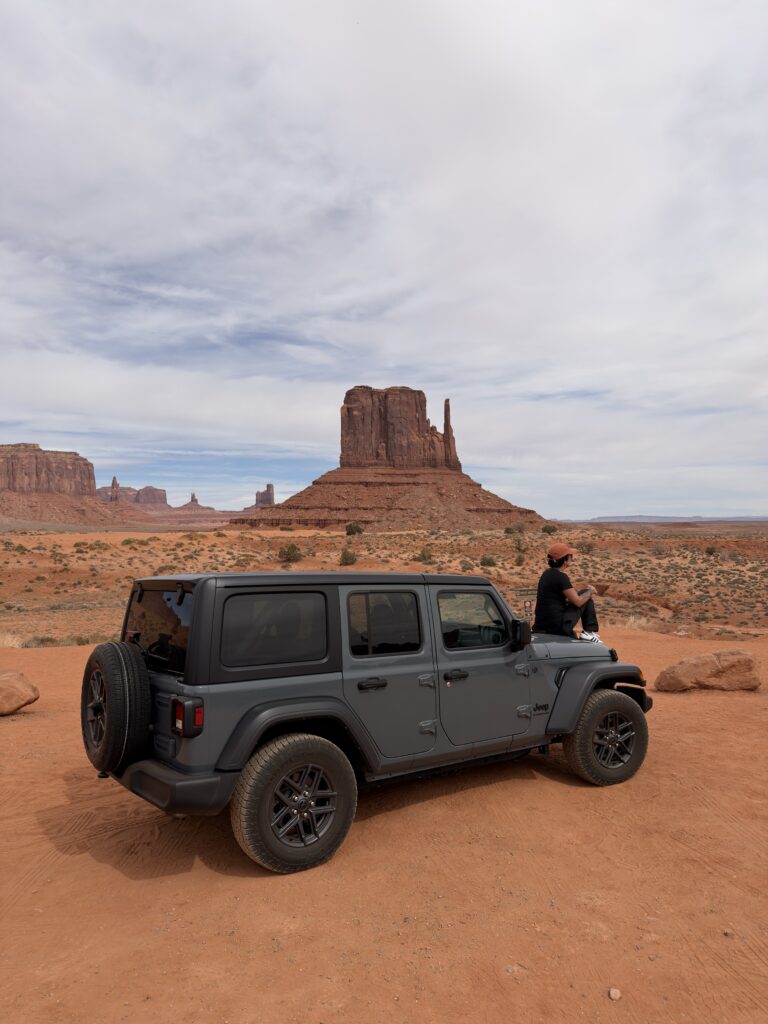 Grauer Jeep Wrangler auf rotem Sandboden im Monument Valley, im Hintergrund der markante West Mitten Butte unter bewölktem Himmel, Person lehnt an der Motorhaube