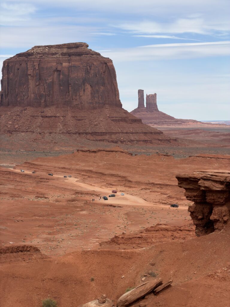 Erhöhter Blick auf den staubigen Valley Drive im Monument Valley mit Fahrzeugen auf der Schotterpiste, links der massige Merrick Butte, dahinter schlanke Felsnadeln.