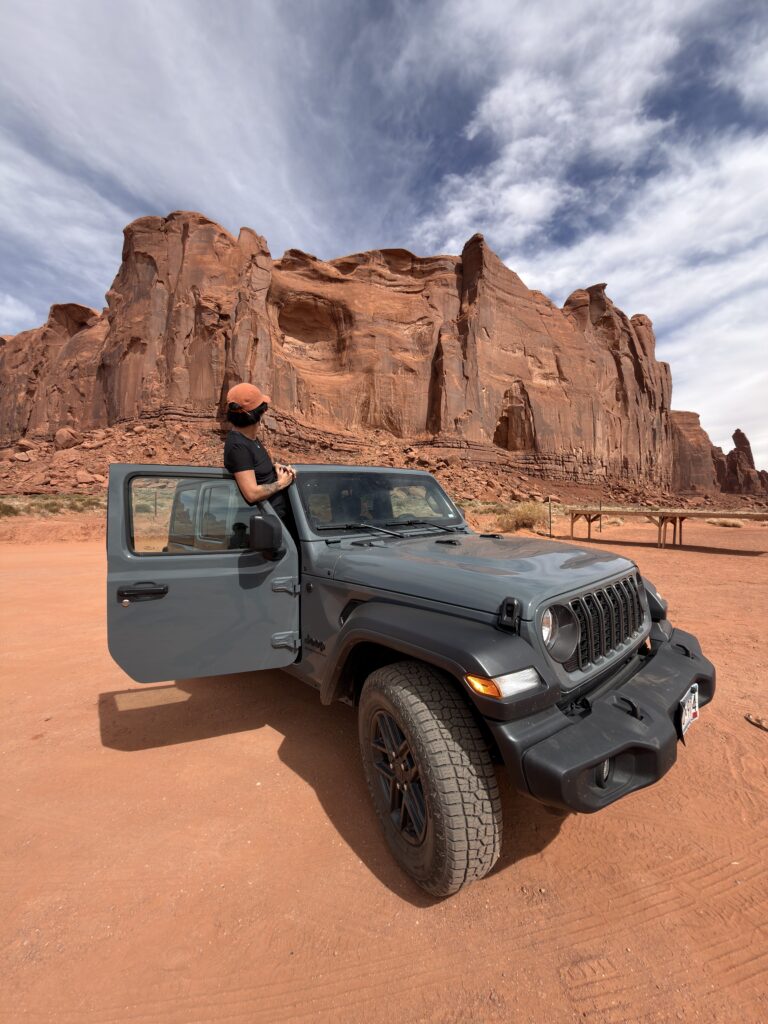 Frau mit orangefarbener Kappe lehnt an der offenen Fahrertür eines grauen Jeep Wrangler im Monument Valley, im Hintergrund mächtige rote Sandsteinwände unter blauem Himmel