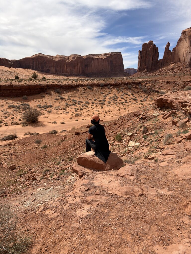 Person mit orangem Cap sitzt von hinten auf einem Sandsteinfelsen im Monument Valley und blickt auf rote Felsformationen und Sandsteinwände.