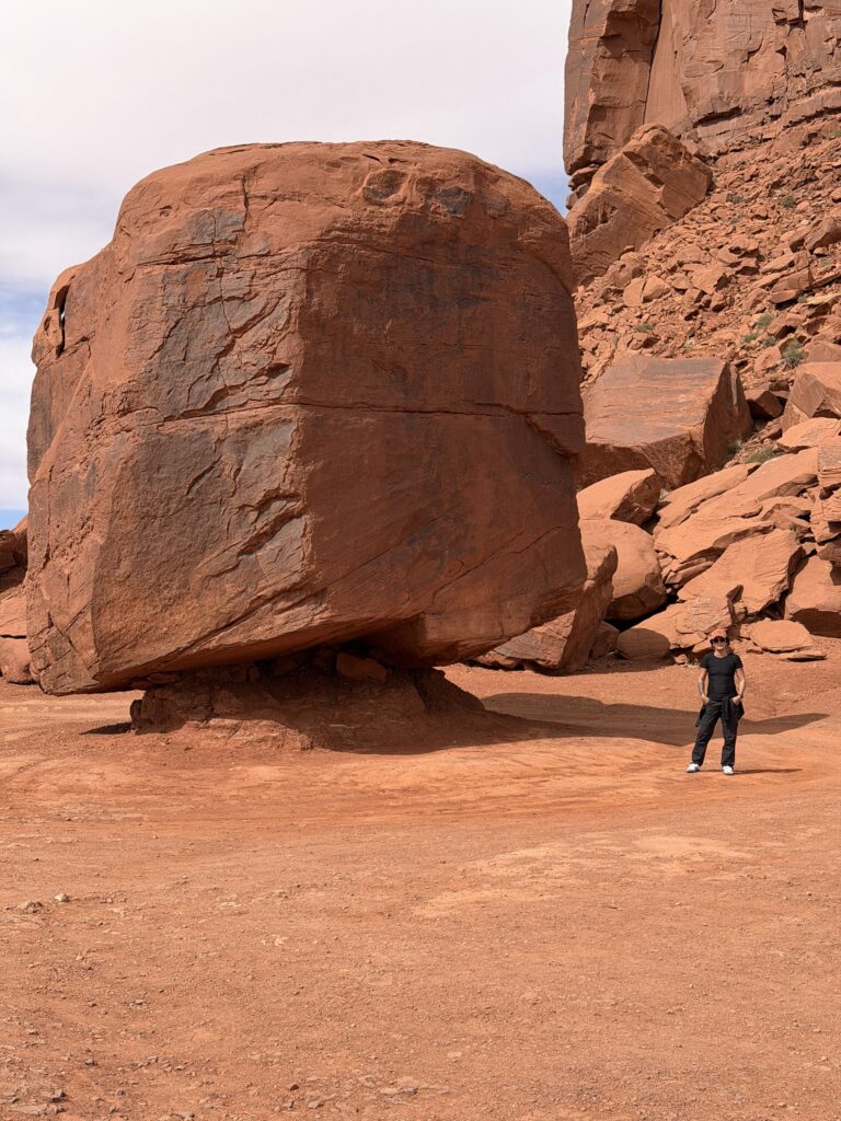 Frau in Schwarz steht neben einem übergroßen abgebrochenen roten Sandsteinblock im Monument Valley, der Größenunterschied ist beeindruckend.