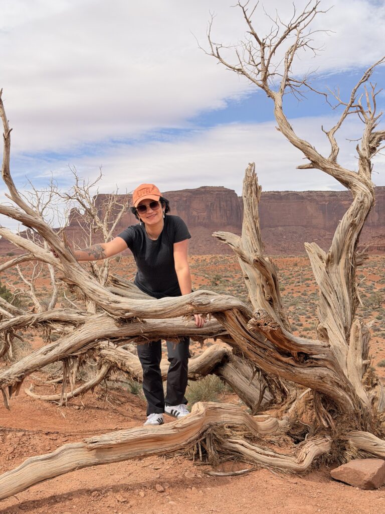 Frau mit orangem Cap lächelt an einem großen silberweißen verwitterten Wacholderbaum im Monument Valley, hinter ihr roter Sand und ein Butte.
