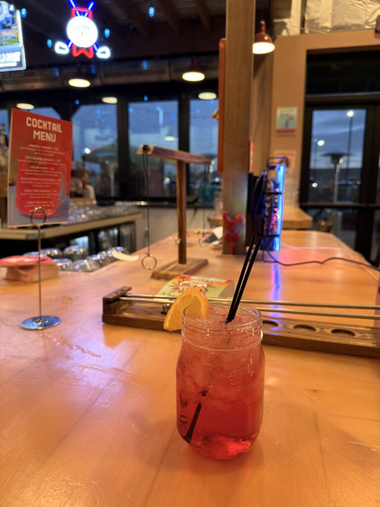 Roter Cocktail im Mason-Glas mit Orangenscheibe und schwarzem Strohhalm auf einer Holzbar, im Hintergrund die Cocktailkarte und Neonlichter.