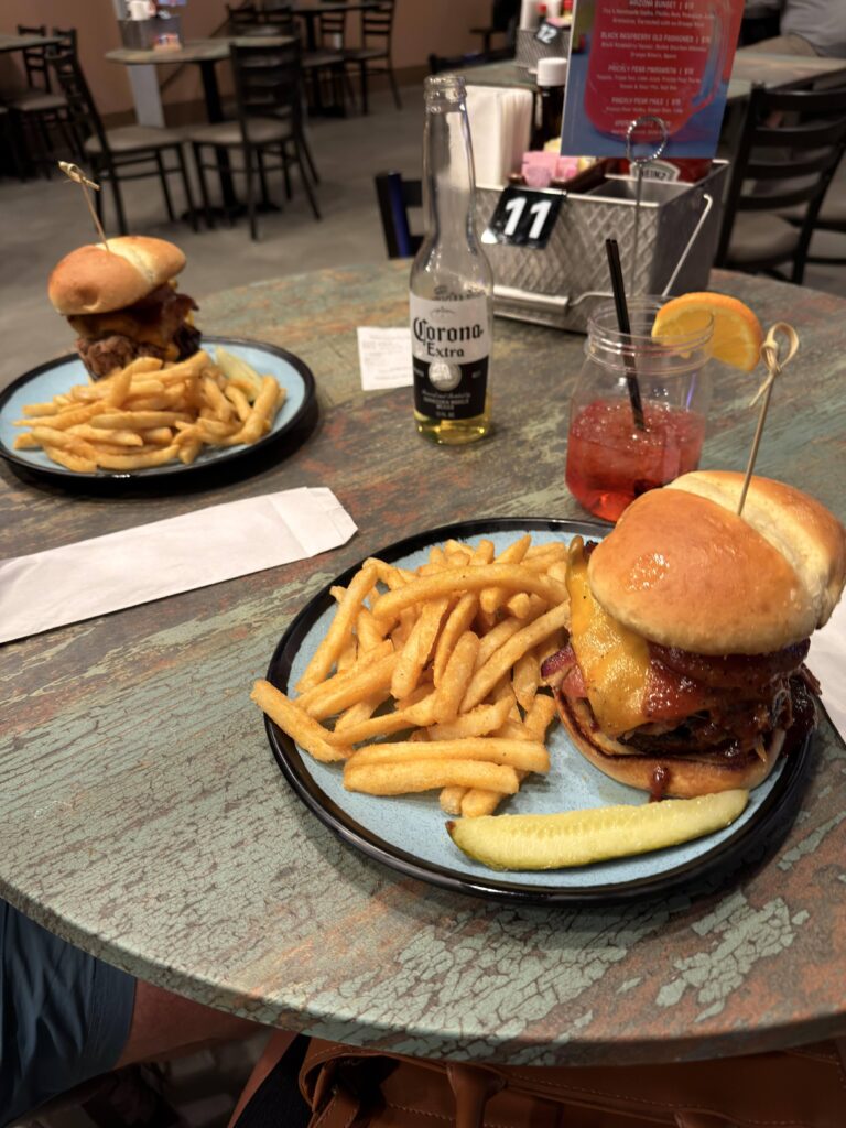 Zwei Teller mit Big Tex Burger und Pommes frites auf rustikalem Tisch, dazu Corona-Bier und roter Cocktail im Mason-Glas.
