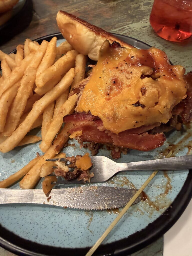 Angeschnittener Big Tex Burger mit geschmolzenem Cheddar, Bacon und Pulled Pork auf einem blauen Teller mit Pommes und Gabel.