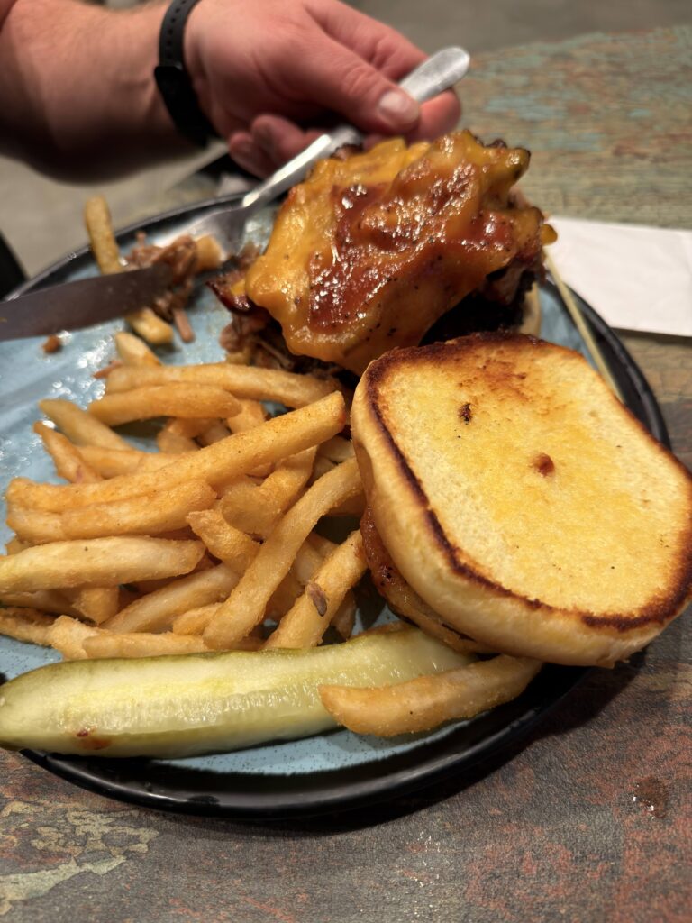 Detailaufnahme des Big Tex Burgers mit geröstetem Bun, geschmolzenem Cheddar, Bacon und Pulled Pork auf blauem Teller mit Pommes und Pickle.