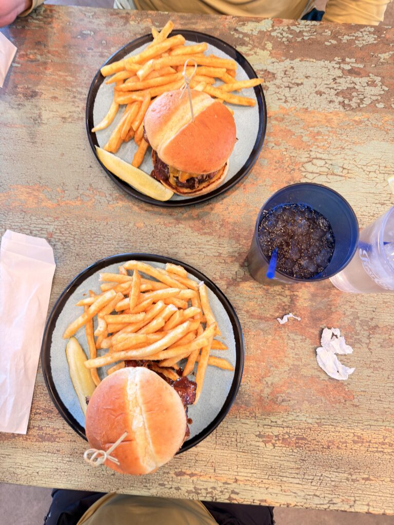 Draufsicht auf zwei Big Tex Burger mit Pommes frites auf rustikalem Tisch von oben, dazu ein Glas Cola.