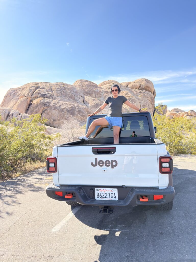 Frau sitzt auf der Ladefläche eines weißen Jeep Gladiator im Joshua Tree National Park, im Hintergrund runde Granitfelsen und blauer Himmel