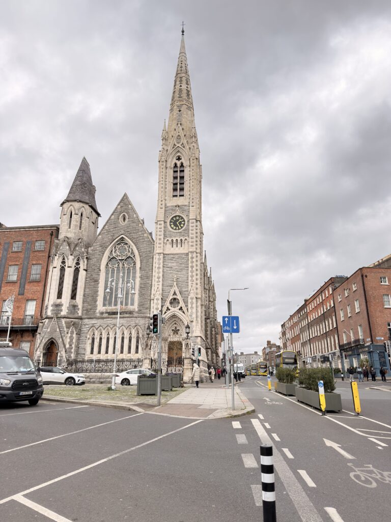 Neugotische Kirche mit hohem Spitzturm und Uhr an einer Hauptstraße in Dublin