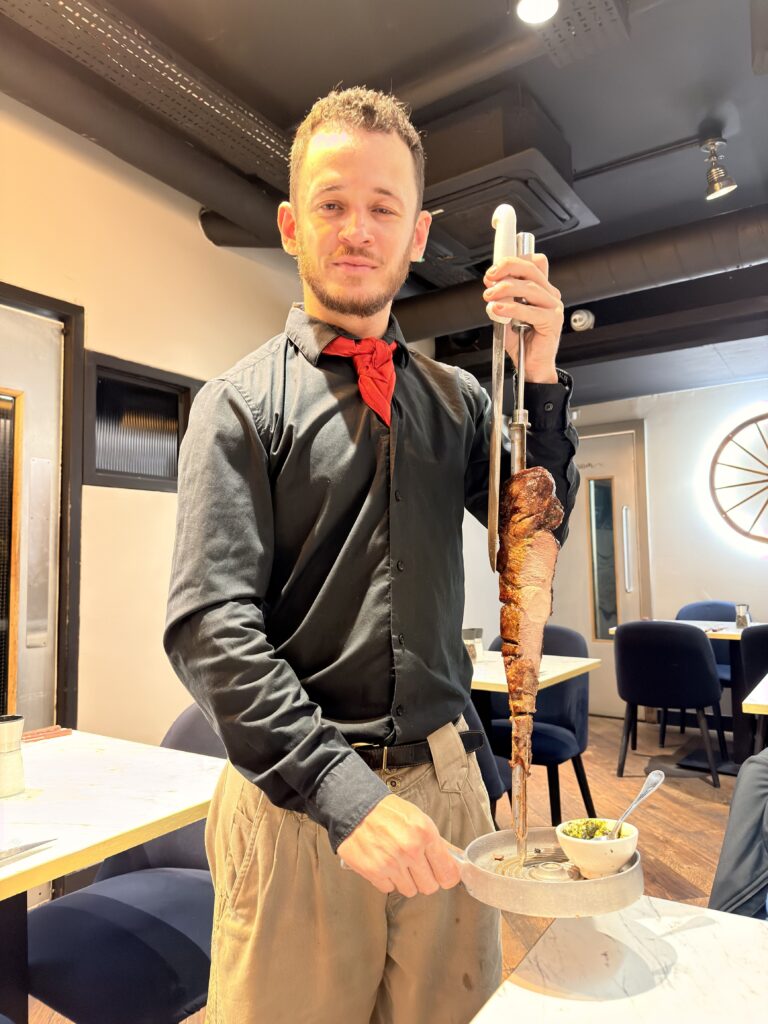 Kellner in schwarzer Uniform präsentiert großen Fleischspieß am Tisch im brasilianischen Restaurant Dublin