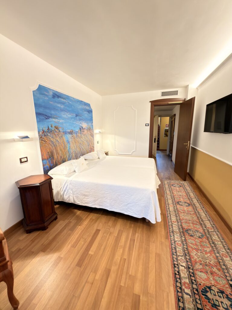 Helles Doppelzimmer im Hotel Villa Mandrina am Gardasee mit Parkettboden, orientalischem Teppich und einem Wandgemälde mit Seemotiv