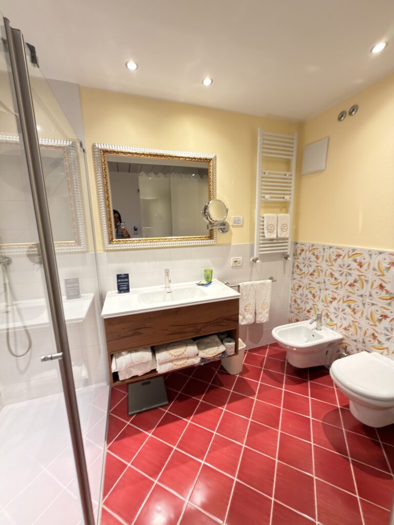 Hochwertiges Badezimmer im Hotel Villa Mandrina Garda mit roten Bodenfliesen, goldgerahmtem Spiegel, Dusche und Bidet
