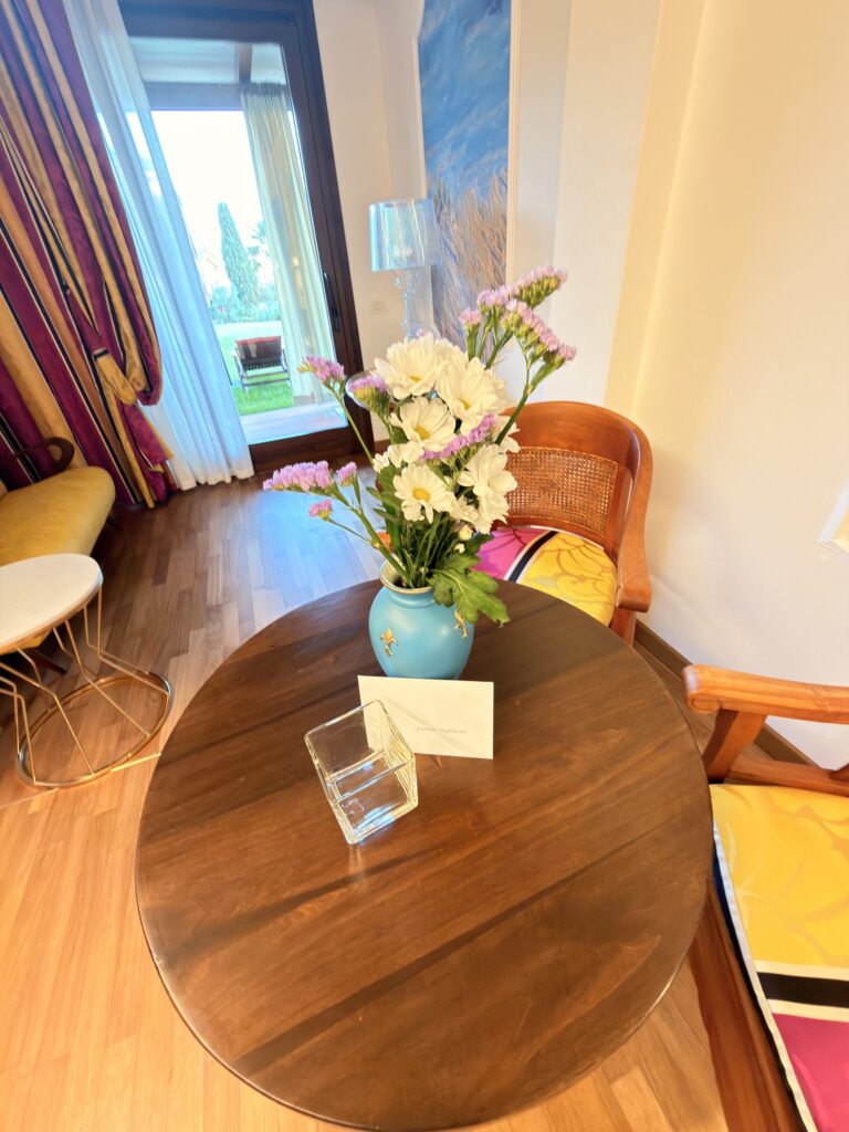 Gemütlicher Wohnbereich eines Hotelzimmers im Hotel Villa Mandrina am Gardasee mit rundem Holztisch, frischen Blumen und Zugang zur Terrasse