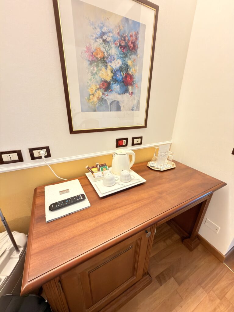 Holzkommode im Hotelzimmer des Hotel Villa Mandrina Garda mit Wasserkocher, Teeservice, Fernbedienung und Blumengemälde an der Wand