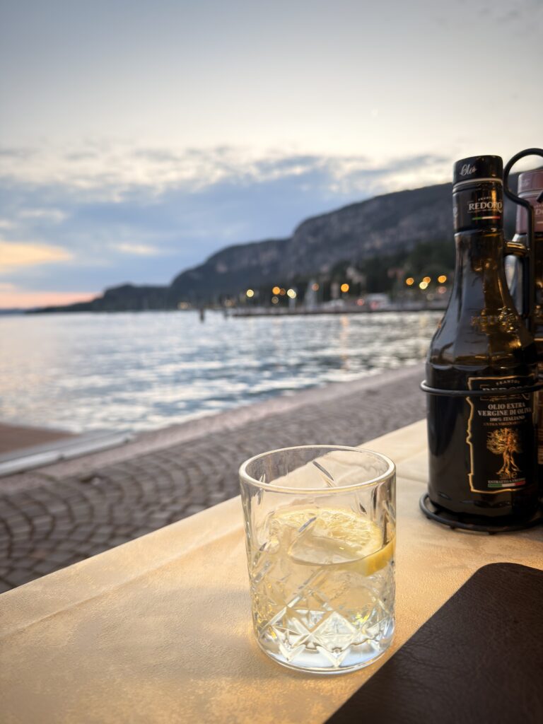 Kristallglas mit Eiswürfeln und Zitronenscheibe auf einem Restauranttisch direkt am Gardasee im El Refol in Garda, im Hintergrund die Uferpromenade mit Lichtern und die Rocca di Garda im Abenddämmer