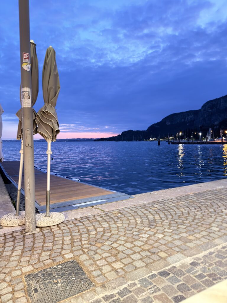 Zusammengefaltete Sonnenschirme auf einem Holzsteg an der Uferpromenade von Garda in der blauen Stunde nach Sonnenuntergang, der Himmel leuchtet in tiefem Blau und Rosa, die Lichter der Promenade spiegeln sich im Wasser
