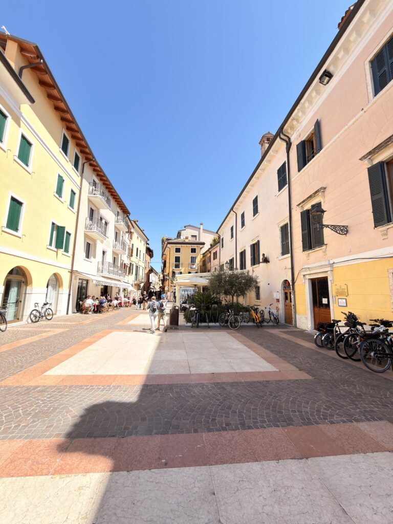 Breite gepflasterte Fußgängerzone in Bardolino mit ockergelben und rosafarbenen Häuserfassaden, Cafés mit Außenbestuhlung und abgestellten Fahrrädern unter strahlend blauem Himmel