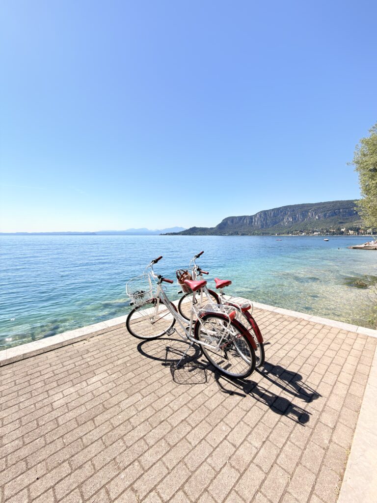 Zwei weiß-rote Citybikes stehen auf einer gepflasterten Uferpromenade direkt am türkisblauen Gardasee, im Hintergrund Felsklippen und Berge