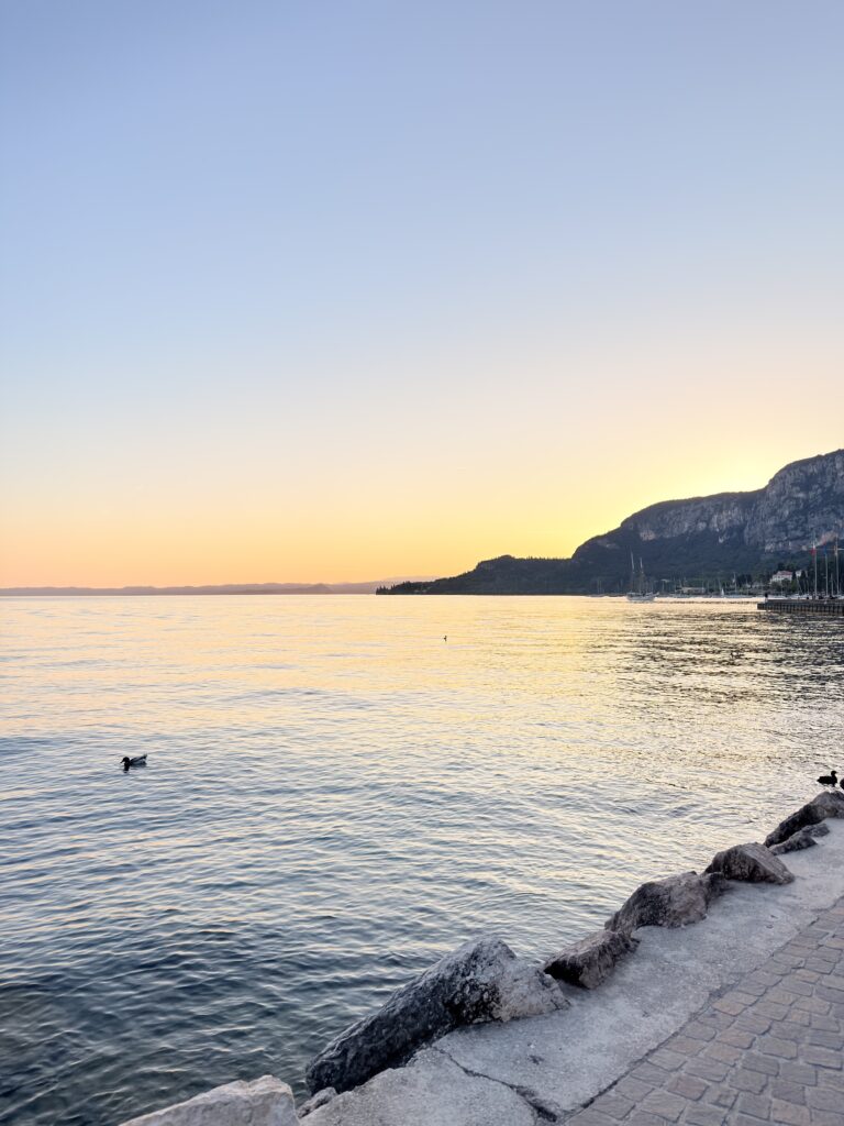 Goldener Sonnenuntergang über dem Gardasee, aufgenommen vom Ufer in Garda mit warmen Orange- und Rottönen am Himmel