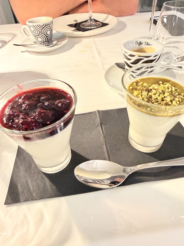 Zwei Gläser Panna Cotta im Hotel Ristorante Giardinetto – links mit dunklem Beerencompot, rechts mit Pistazien-Topping, dazu zwei Espressotassen im Hintergrund
