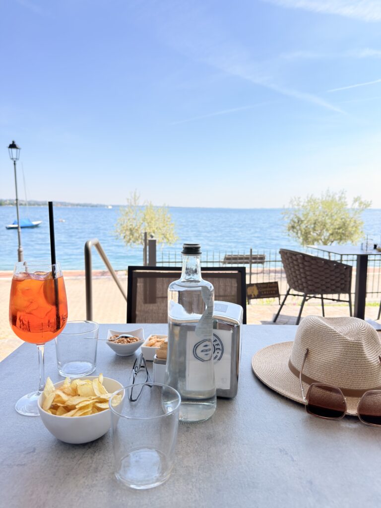 Aperol Spritz, Wasserflasche, Chips und Strohhut auf einem Tisch der Außenterrasse des Bistro du Lac in Garda, direkter Blick auf den blauen Gardasee mit Strand und Olivenbäumen