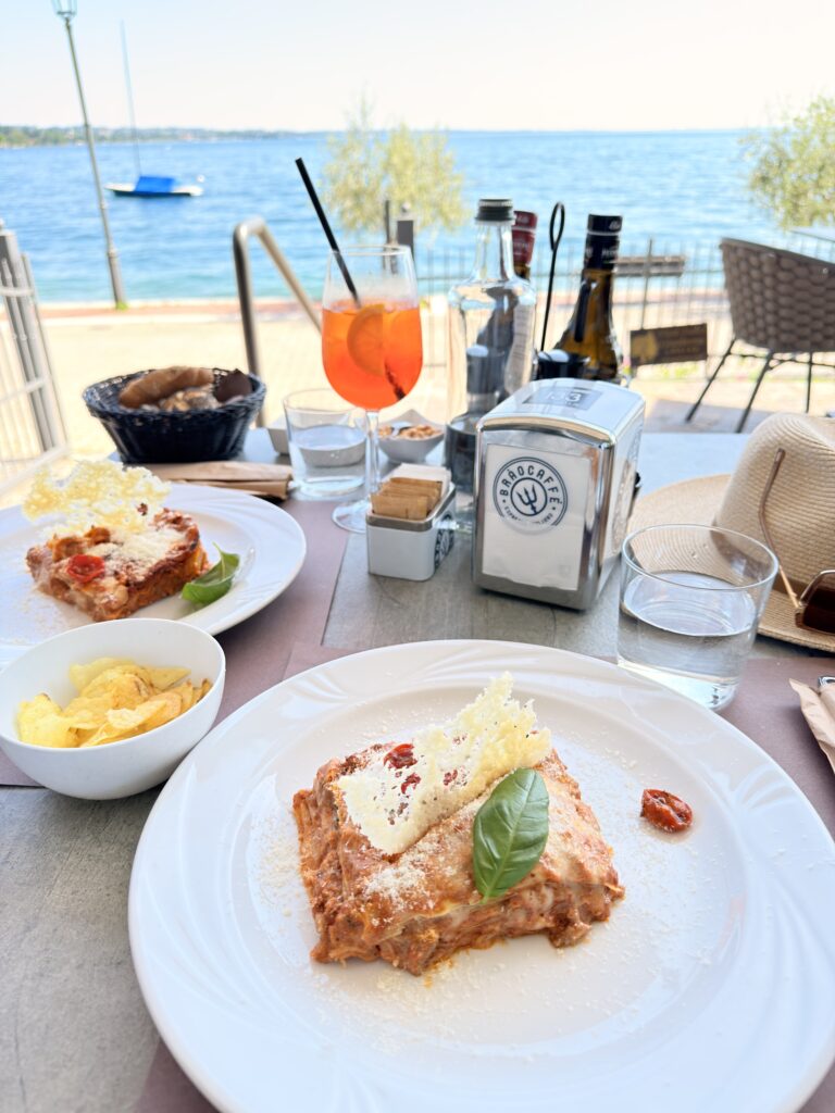 Teller mit Lasagne, garniert mit Basilikum und Parmesancracker, auf einem Restauranttisch direkt am Gardasee im Bistro du Lac in Garda, im Hintergrund ein Aperol Spritz und der blaue See