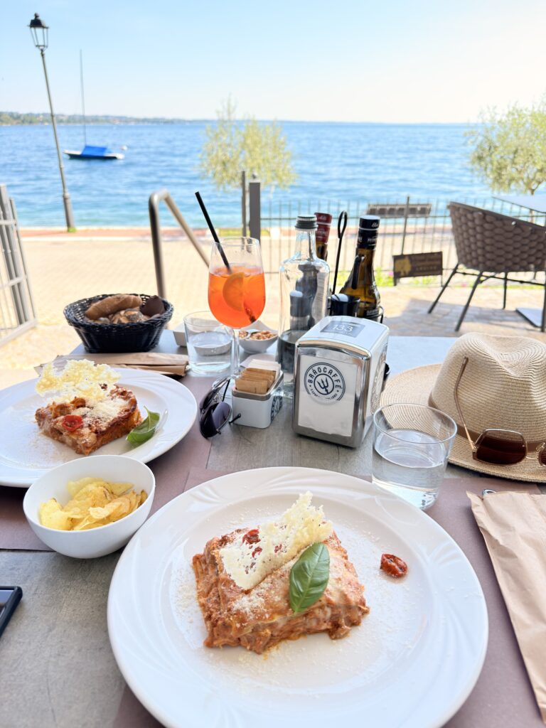 Gedeckter Mittagstisch auf der Seeterrasse des Bistro du Lac in Garda mit zwei Tellern Lasagne, einem Aperol Spritz, Brotkorb, Chips und Strohhüten, im Hintergrund der blaue Gardasee