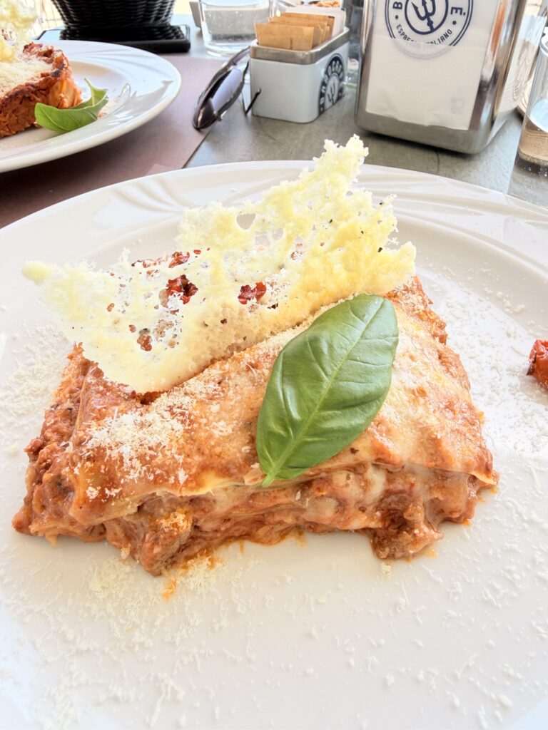 Nahaufnahme einer goldbraun gebackenen Lasagne al Forno mit knusprigem Parmesancracker und frischem Basilikumblatt auf weißem Teller, serviert im Bistro du Lac in Garda am Gardasee