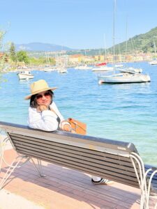 Ich mit Strohhut und Sonnenbrille sitzt entspannt auf einer Bank an der Uferpromenade im Norden von Garda, im Hintergrund Segelboote und die bunte Häuserzeile am gegenüberliegenden Ufer