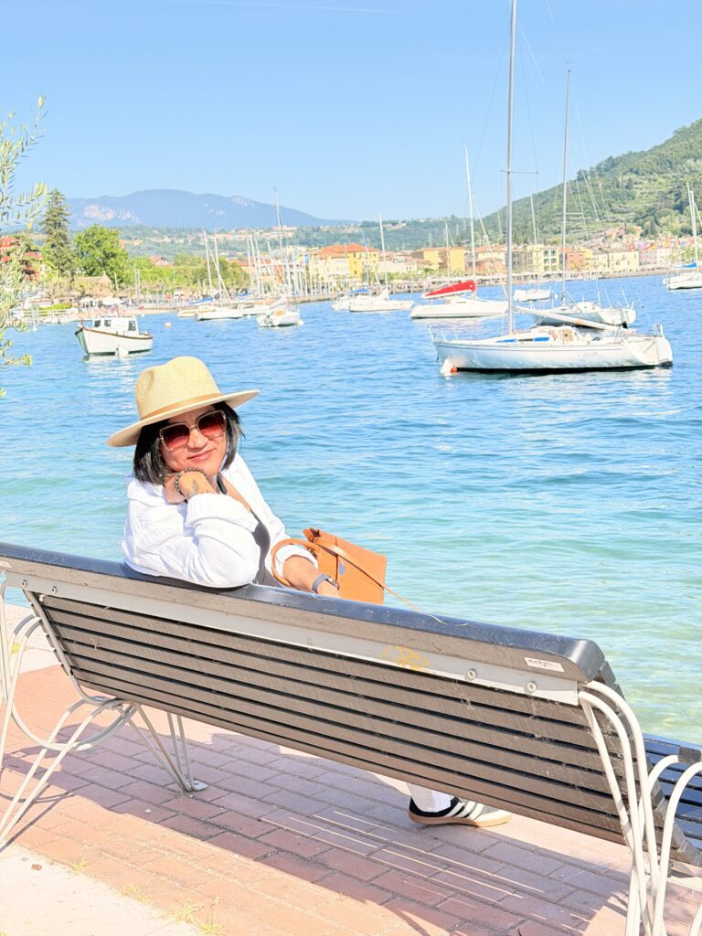 Ich mit Strohhut und Sonnenbrille sitzt entspannt auf einer Bank an der Uferpromenade im Norden von Garda, im Hintergrund Segelboote und die bunte Häuserzeile am gegenüberliegenden Ufer