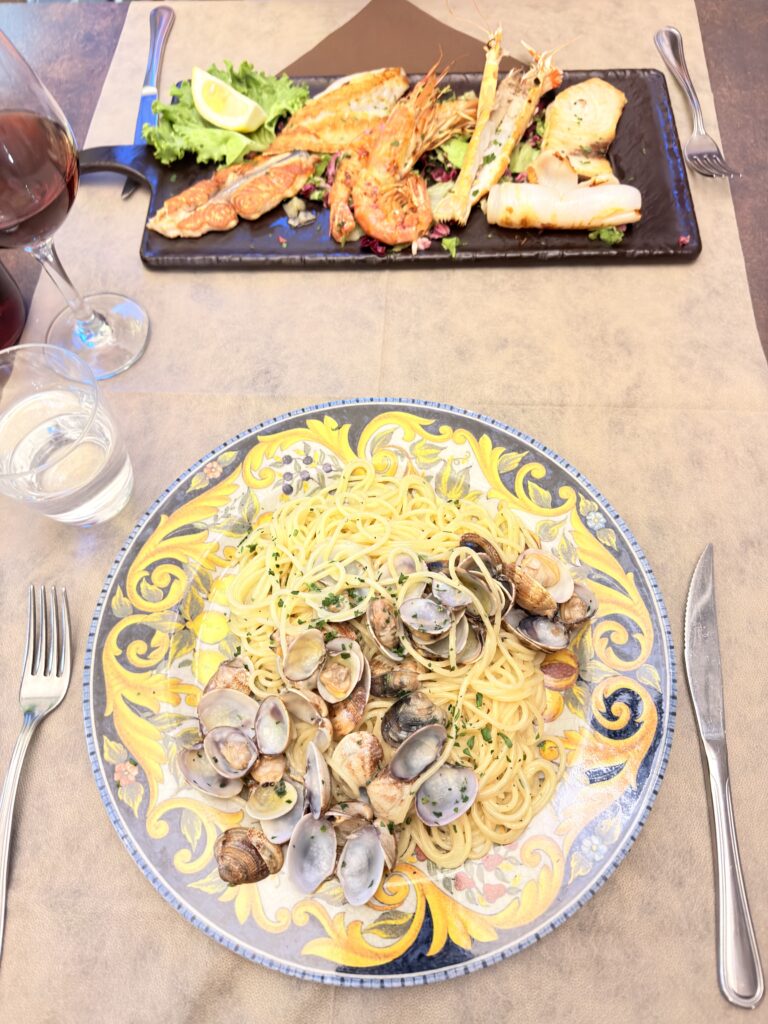 Spaghetti alle Vongole auf einem handbemalten blau-gelben Keramikteller im Vordergrund, dahinter ein schwarzer Schieferteller mit gegrillten Scampi und Fisch sowie einem Glas Rotwein, serviert im Restaurant Bussola in Garda