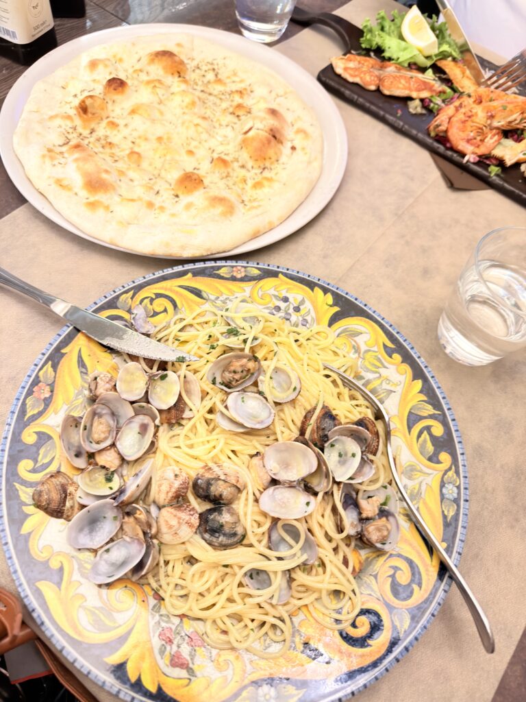 Tischansicht im Restaurant Bussola in Garda mit Spaghetti alle Vongole auf Keramikteller, einer goldbraun gebackenen Focaccia und einem Schieferteller mit gegrillten Meeresfrüchten