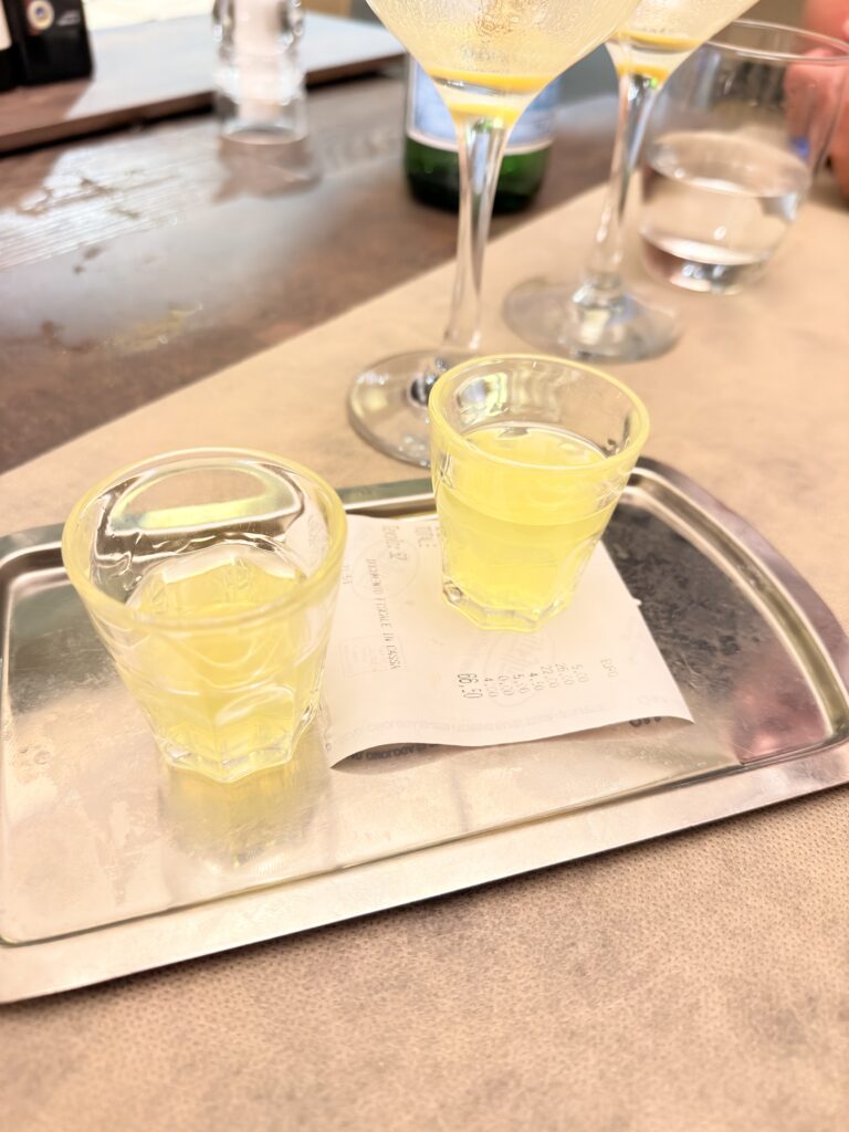 Zwei kleine Gläser mit leuchtend gelbem Limoncello auf einem silbernen Tablett mit der Rechnung, im Hintergrund zwei Sektgläser, serviert als Digestivo im Restaurant Bussola in Garda