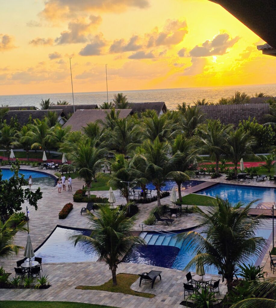 Blick vom Balkon der Ferienwohnung auf Pools, Palmen und einen goldgelben Sonnenuntergang über dem Meer