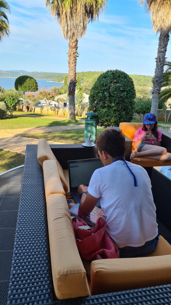 Frau arbeitet am Laptop auf einer Poolterrasse unter Palmen mit Blick auf die Bucht, im Hintergrund eine weitere Person mit Handy