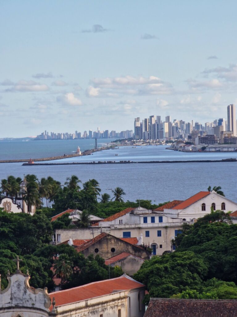 Blick von Olinda über die Bucht auf die Skyline von Recife im Abendlicht, im Vordergrund Kolonialdächer und Palmen