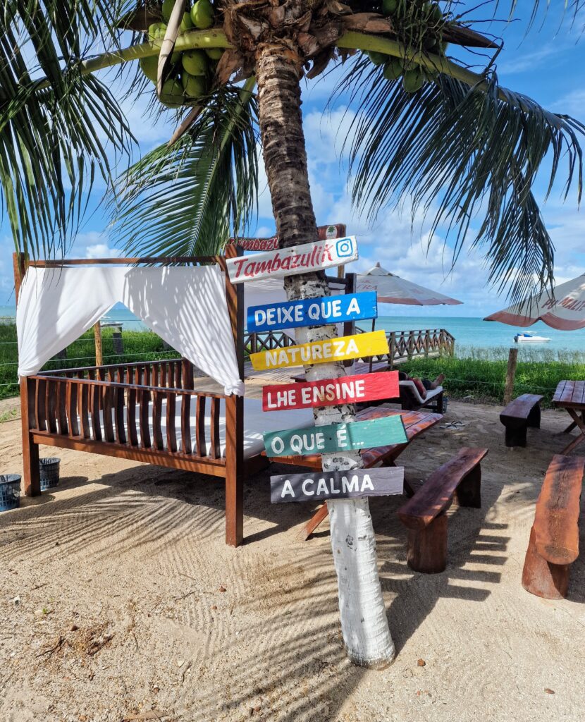 Bunte Wegweiser-Schilder mit Sprüchen an einer Kokospalme vor dem Strandbett des Tambazulik Beach Club in Maragogi