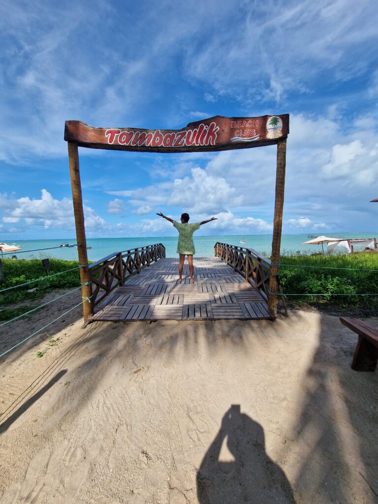 Frau mit ausgebreiteten Armen auf einem Holzsteg unter dem Schild "Tambazulik Beach Club" mit Blick aufs türkisfarbene Meer